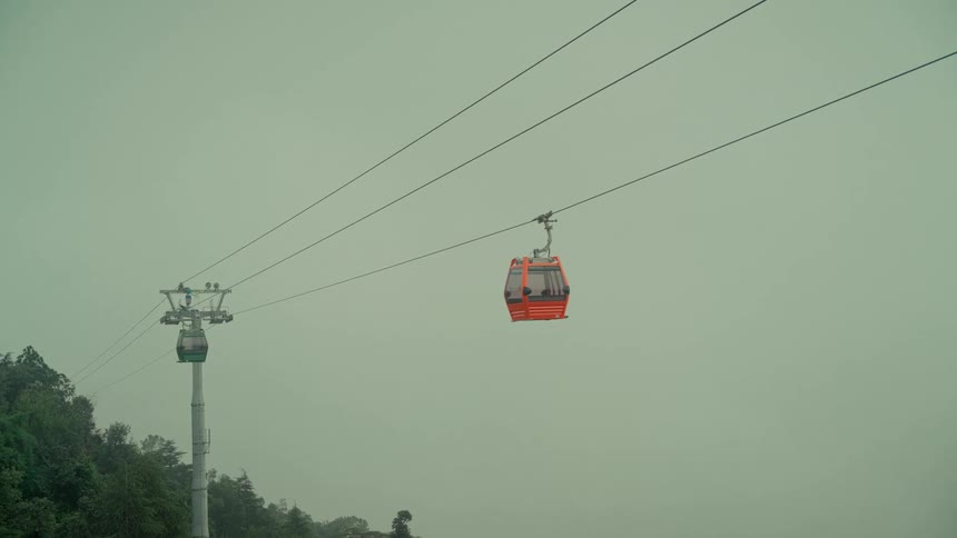 An orange and a green gondola pass each other on a cableway, moving through a thick fog over a mountain forest
