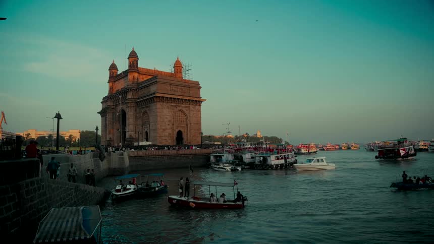The iconic Gateway of India in Mumbai stands over a busy harbor filled with boats during a serene sunset