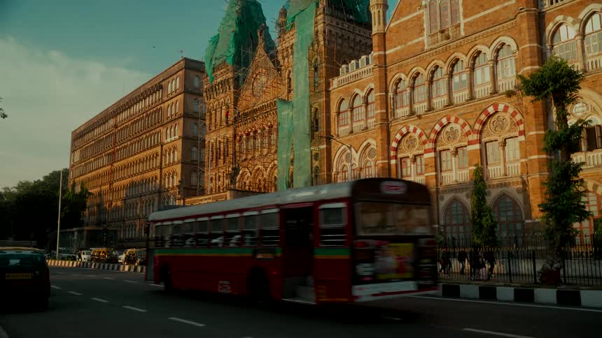 A red city bus drives past the ornate architecture of the Chhatrapati Shivaji Maharaj Terminus in Mumbai during golden hour