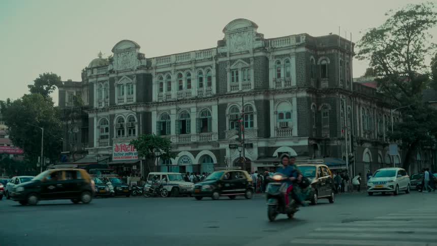 Slow motion shot of a bustling city intersection with cars and scooters passing in front of a historic colonial building in Mumbai, India