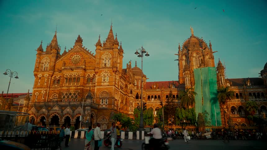 Golden hour sunlight illuminates the magnificent Chhatrapati Shivaji Maharaj Terminus as traffic and pedestrians move through the busy Mumbai streets
