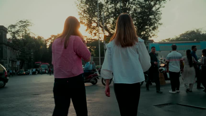 A slow-motion, rear-view shot of two young women walking through a bustling city intersection as the sun sets