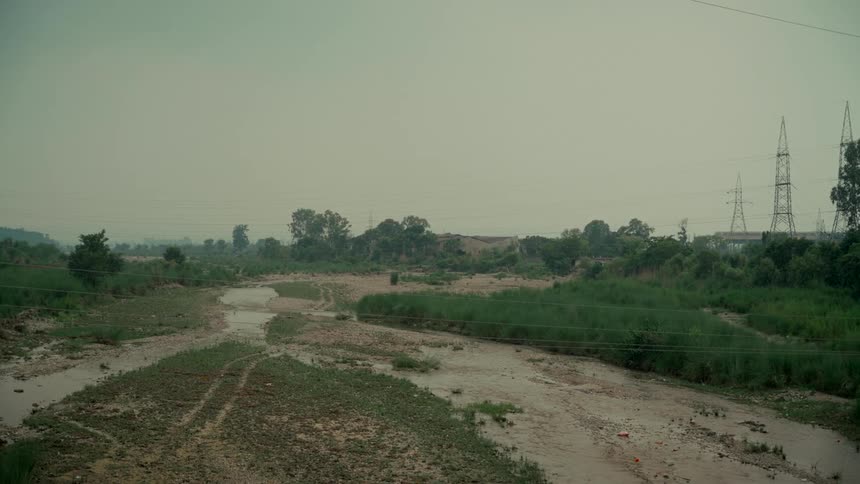 A mostly dry riverbed with a small stream winds through a green landscape under an overcast sky, with large electricity pylons in the background