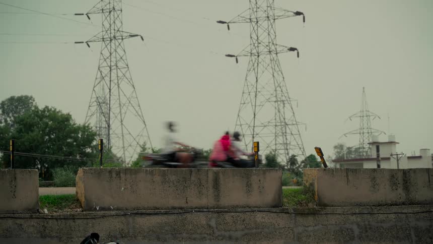 Blurred motorcycles travel along a highway with towering electricity pylons looming in the background under a gray sky