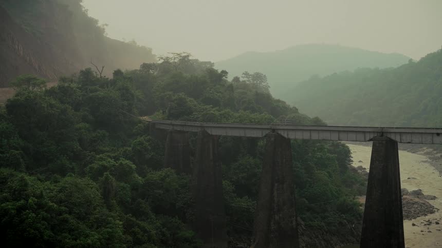 An impressive concrete viaduct stretches across a river in a remote and hazy jungle valley