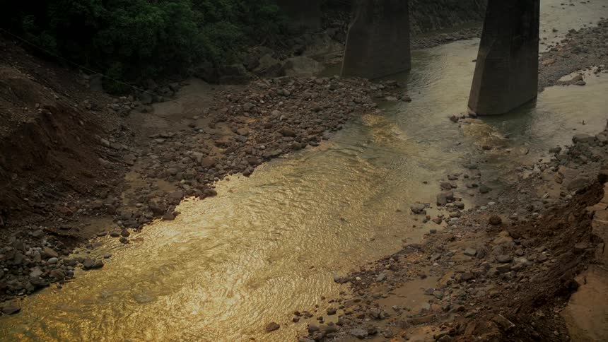 A murky river reflecting golden light flows beneath a bridge, past rocky, eroded banks after a landslide