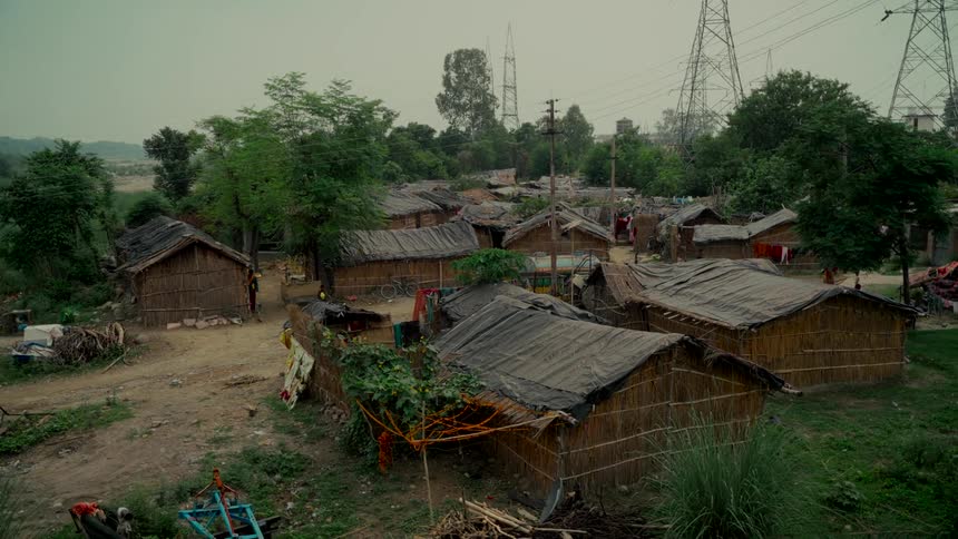 A wide, static shot shows a makeshift village with thatched huts under an overcast sky, with large electrical power lines in the background