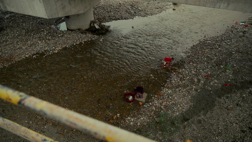 A high-angle view shows people doing laundry in a shallow, rocky river flowing underneath a concrete bridge