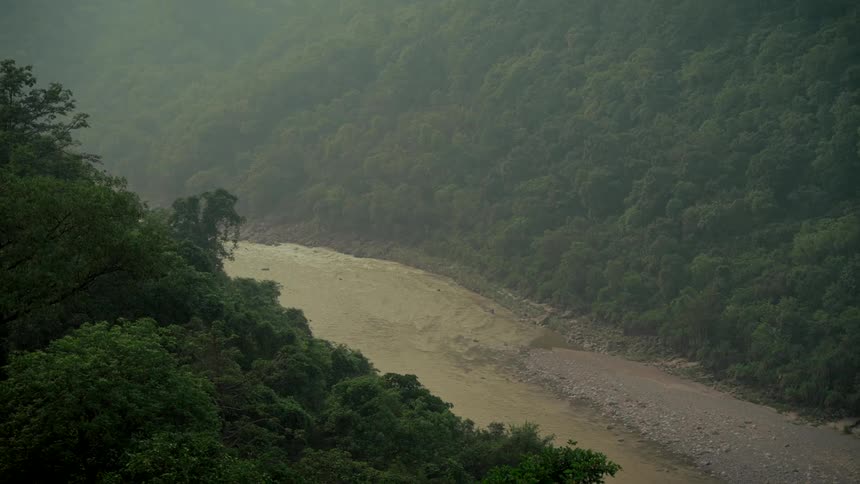 A wide, muddy river flows through a dense, misty jungle valley during a rainstorm