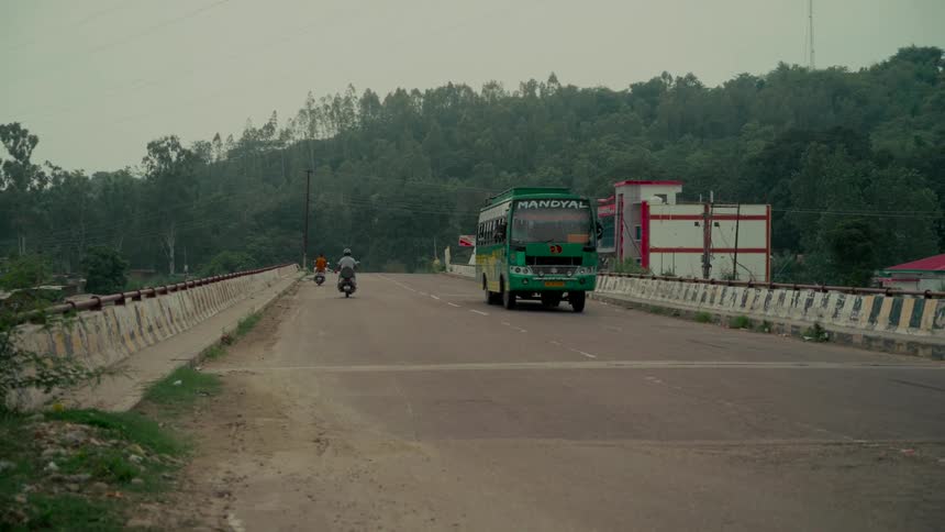 A green public bus and several scooters travel along a bridge in a rural Indian landscape on an overcast day