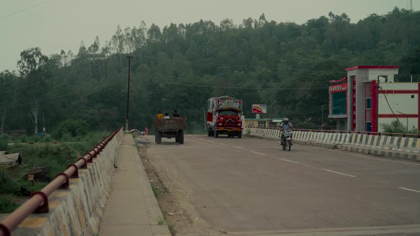A tractor and decorated truck drive across a rural bridge in India with forested hills in the background