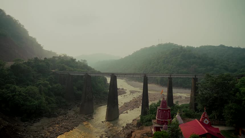A scenic wide shot of an old bridge spanning a river in a lush green valley with a Hindu temple in the foreground on a hazy day