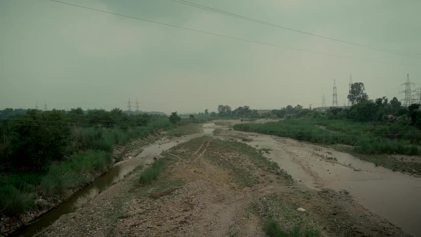 A wide, static shot of a shallow, muddy river winding through a desolate rural landscape with power lines overhead