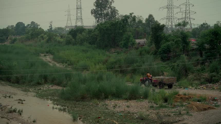 A farmer drives a tractor down a dirt path in a rural village under towering electricity pylons