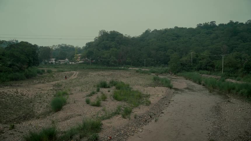 A lone figure in red walks along a vast, rocky riverbed surrounded by dense green forests under a grey sky