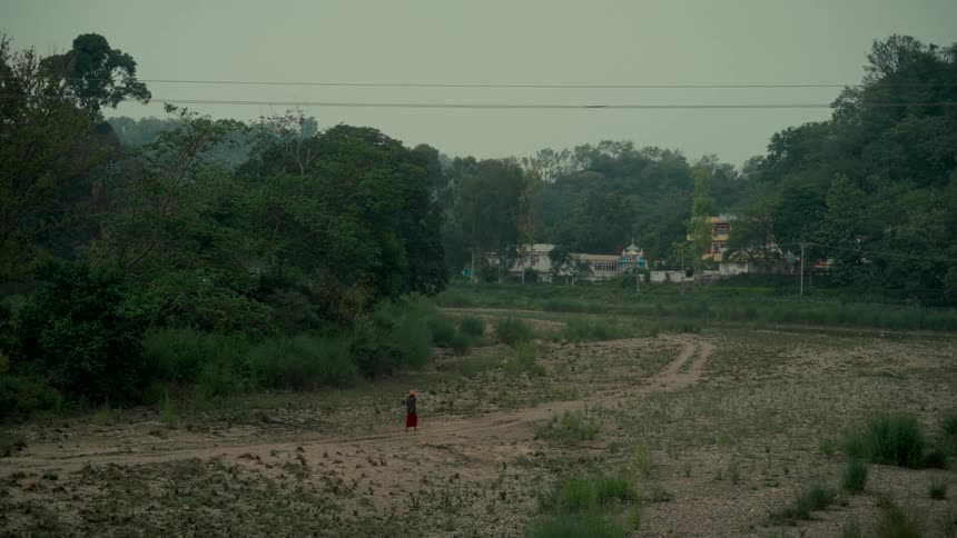 A solitary figure in orange and red robes walks along a dry riverbed path through a vast, green valley
