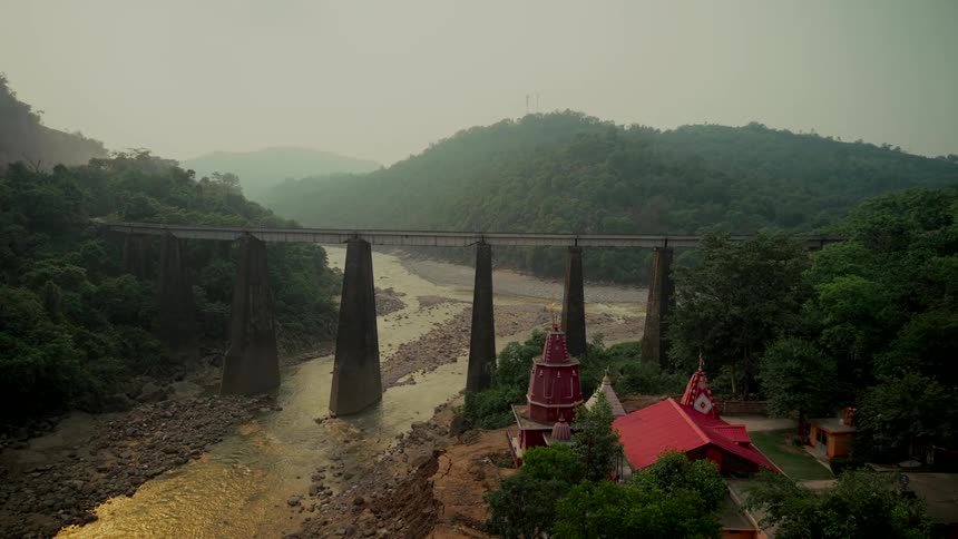 A serene view of a large bridge spanning a river valley in the mountains of India, with a traditional Hindu temple nestled on the riverbank