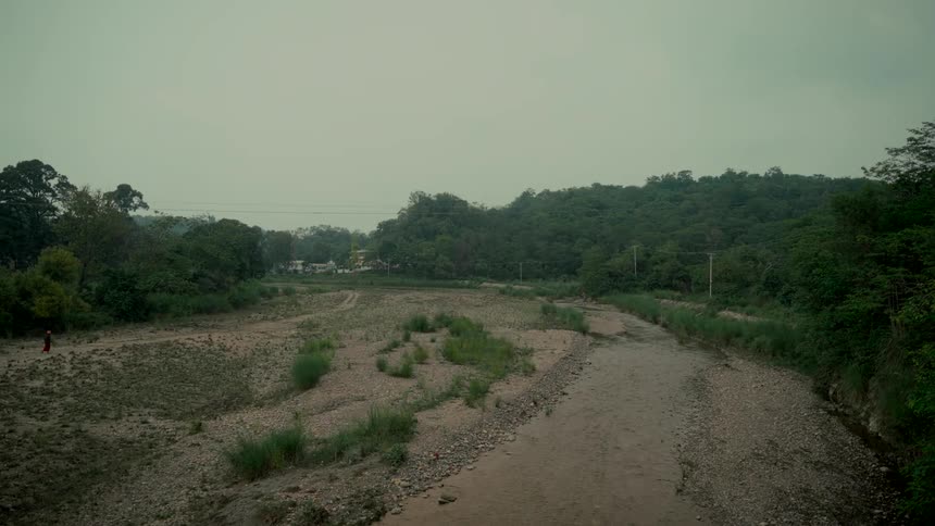 A person in red walks along a path next to a mostly dry riverbed surrounded by a lush green forest on a cloudy day