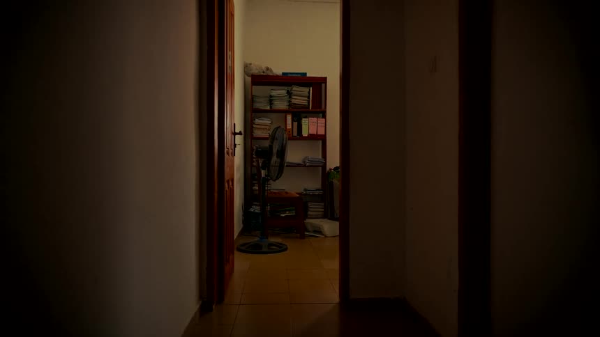 A mysterious static shot from a dark hallway looks into a dimly lit room with a bookshelf and a stationary fan