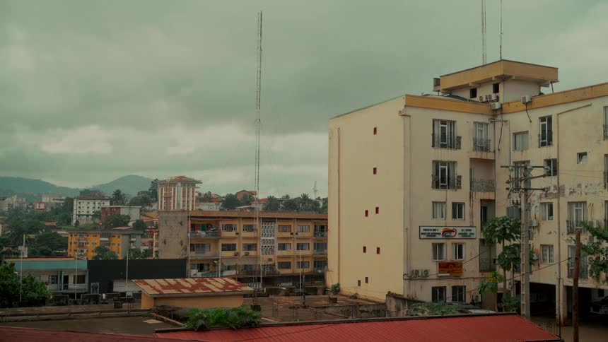 A static high-angle view shows the rooftops and buildings of an African city under a cloudy, overcast sky with rolling hills in the distance