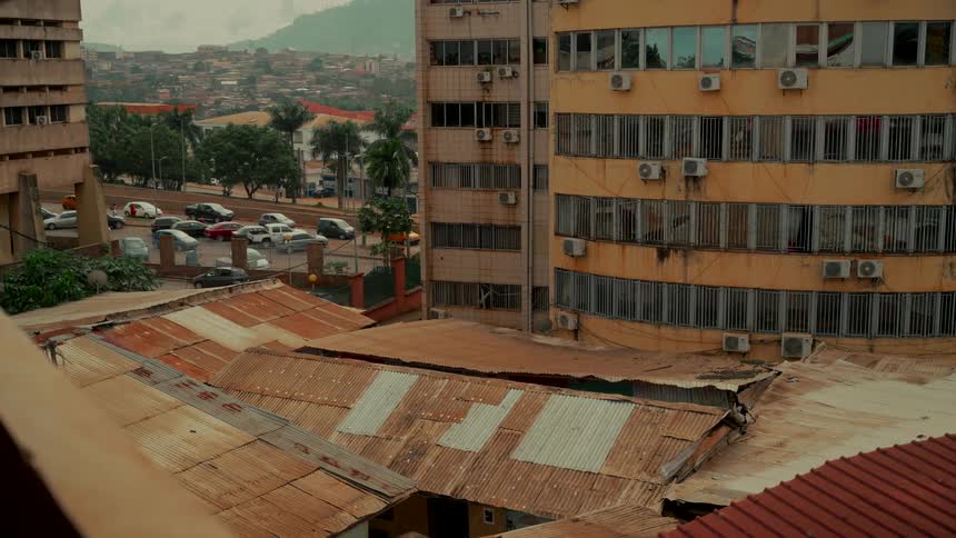 Overlooking the rusty tin roofs of a city, a weathered yellow apartment building stands with traffic passing by in the background