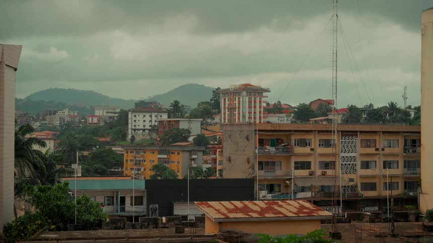 A wide, static view of a city in Africa, showing a mix of architecture against a backdrop of green hills under a cloudy sky