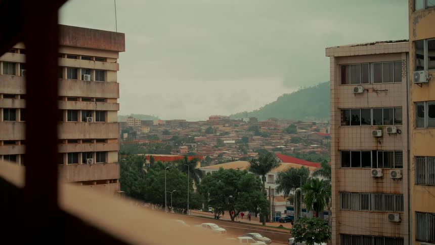 A wide, observational view of a city nestled in green hills, framed by weathered buildings on a cloudy day
