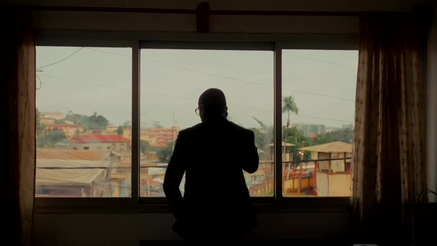 A silhouetted man in a suit makes a phone call while looking out of a window over a city