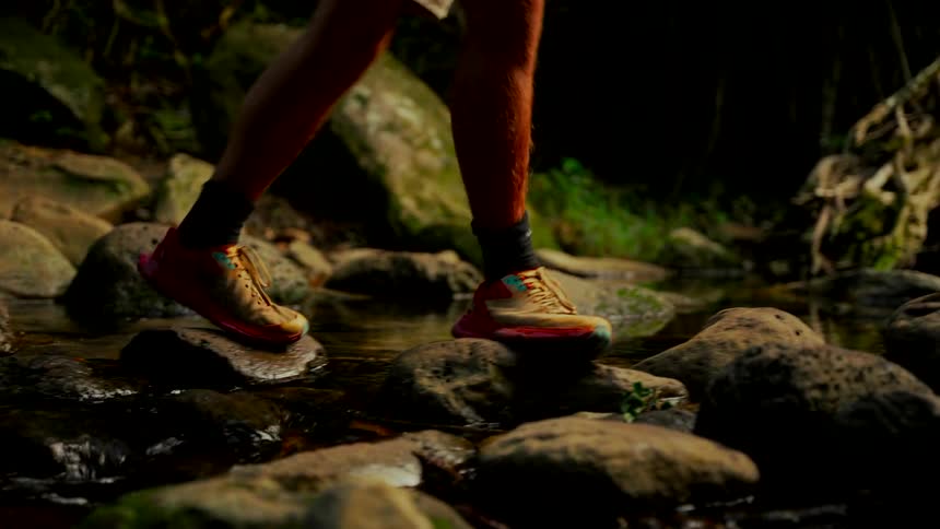 A low angle shot of a hiker's feet in athletic shoes carefully crossing a rocky stream in a dark forest