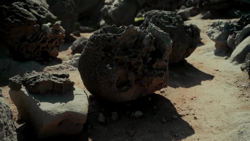 Slow pullback shot reveals a desolate landscape of dark, porous lava rocks resting on sandy terrain