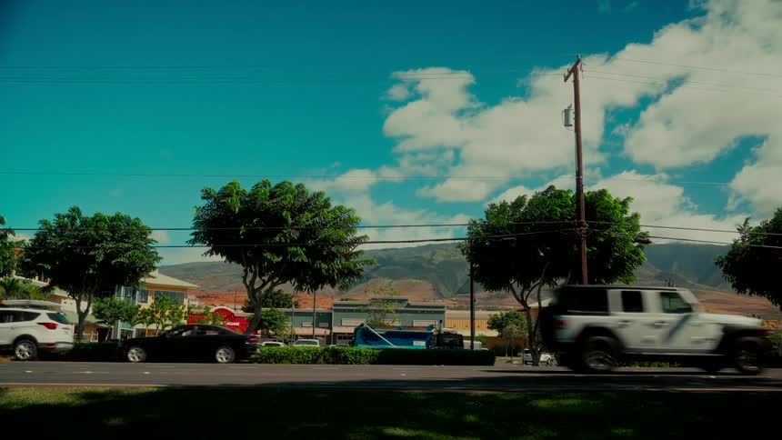 Traffic flows down a sunny street in a tropical town with mountains rising in the background under a blue sky