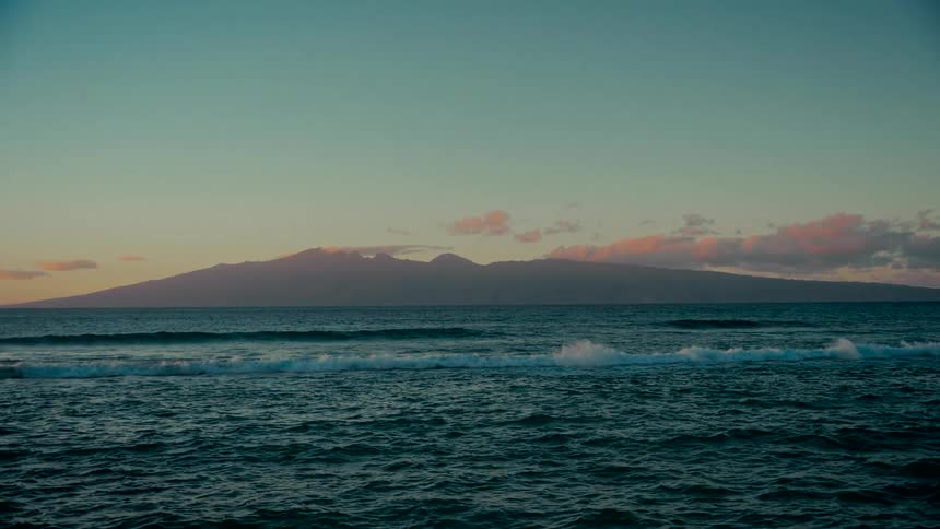 A tranquil and cinematic slow-motion shot captures peaceful ocean waves with a large island on the horizon at dusk