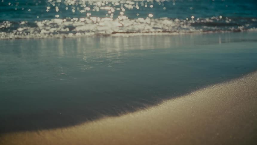 Sparkling sun reflects on the tranquil sea as gentle waves wash ashore in slow motion