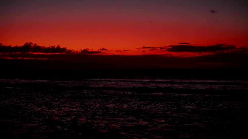 A dramatic deep red sunset casts a silhouette over the dark ocean waves