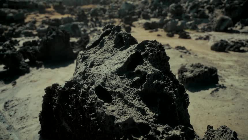 A close-up of a porous, dark lava rock resting on the sandy ground of a desolate landscape