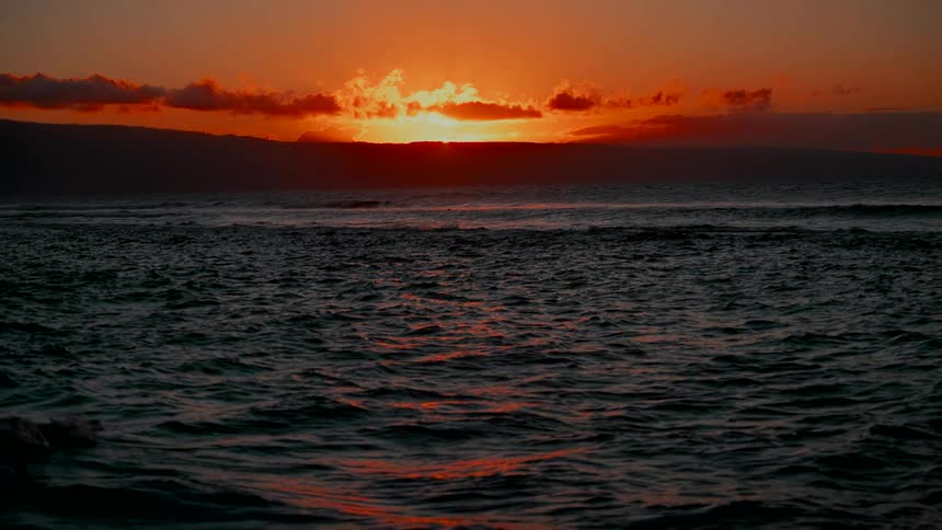 The sun sets behind a distant island, casting a brilliant orange glow over the rolling ocean waves