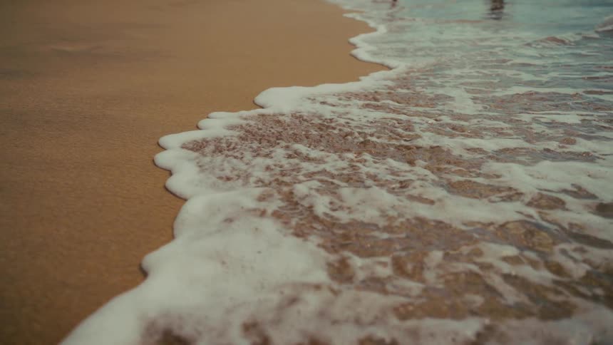 A close-up slow-motion view of a gentle ocean wave's white foam receding from the wet golden sand of a tranquil beach