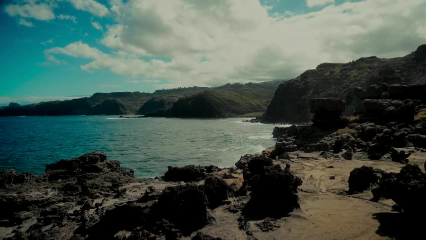 A dramatic slow-motion wide shot of a rocky volcanic coastline with vibrant turquoise water and a cloudy sky