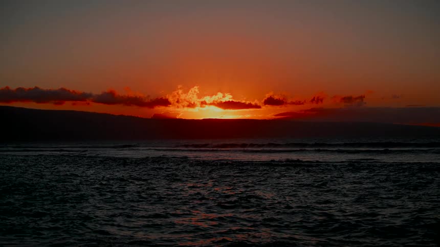 A brilliant orange sun sets behind a silhouetted island, casting its warm glow over the tranquil ocean
