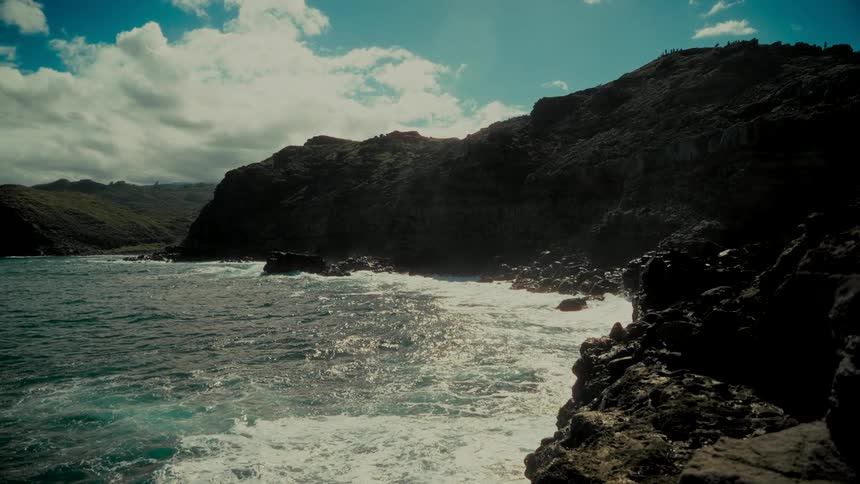 Sunlight reflects off the choppy ocean as waves crash against a dramatic volcanic rock coastline