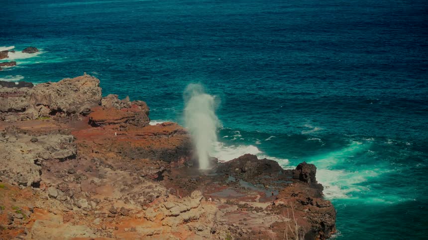 Slow motion shot of a powerful blowhole erupting on a rocky volcanic coast next to a turquoise ocean, creating a small rainbow in the mist