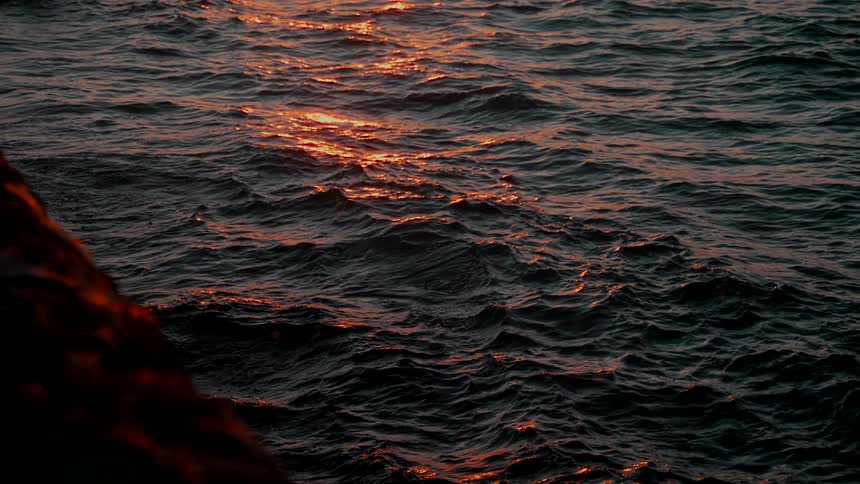 The deep orange glow of the sunset reflects on the dark, mesmerizing ocean waves in slow motion