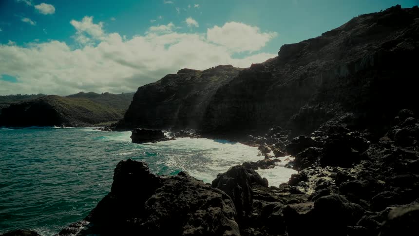 Slow motion waves crash against a dramatic rocky coastline under a bright, partly cloudy sky