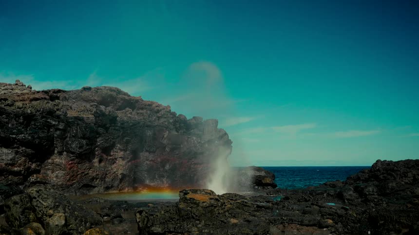 A powerful ocean blowhole erupts in slow motion on a rugged volcanic coastline, creating a rainbow in its misty spray against a bright blue sky