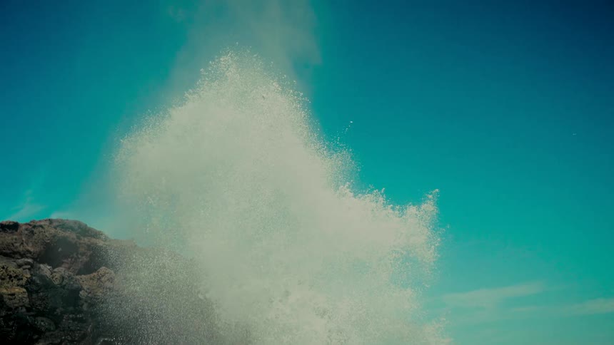 A dramatic slow-motion shot captures a natural ocean blowhole erupting with immense force against a rocky volcanic coast
