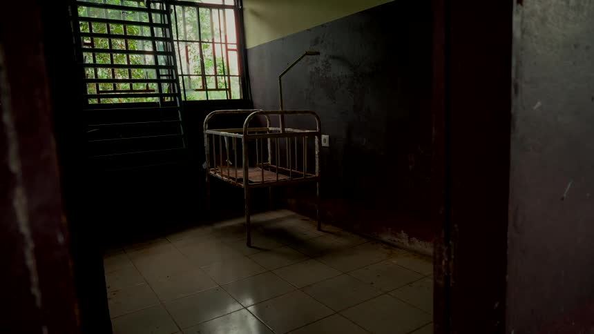 An empty, rusty baby crib sits ominously in the center of a dark, dilapidated room in an abandoned building