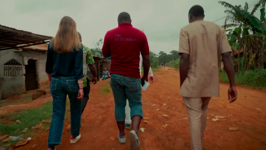 A diverse group of people walk together in slow motion down a red dirt road through a rural village