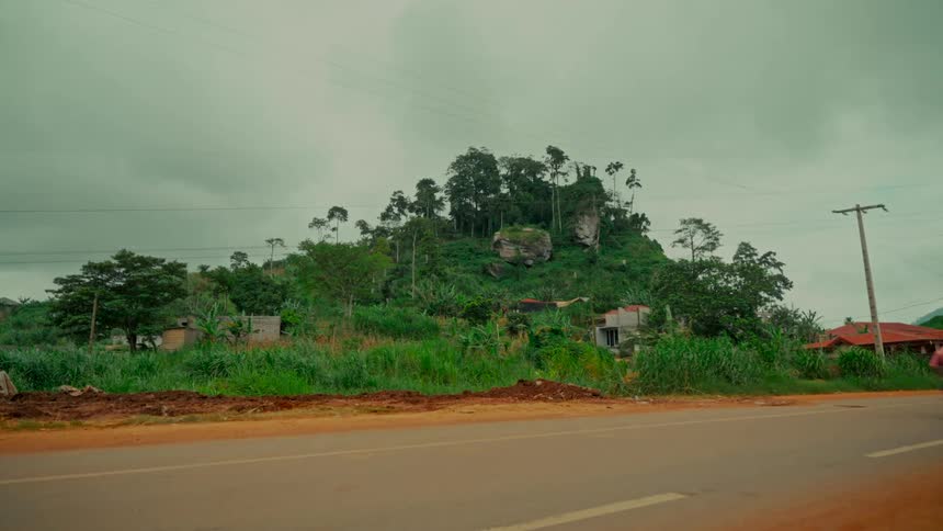A motorcycle with passengers drives on a rural road past a lush, tree-covered hill under a cloudy sky