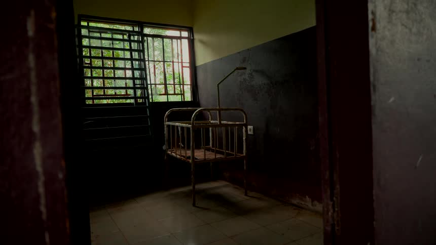 A haunting view of a derelict room with a rusty, empty baby crib next to a barred window