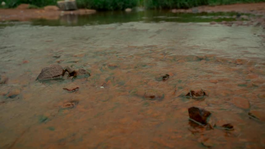 A close-up view of a shallow, calm stream as it gently flows over a reddish-brown rocky bed